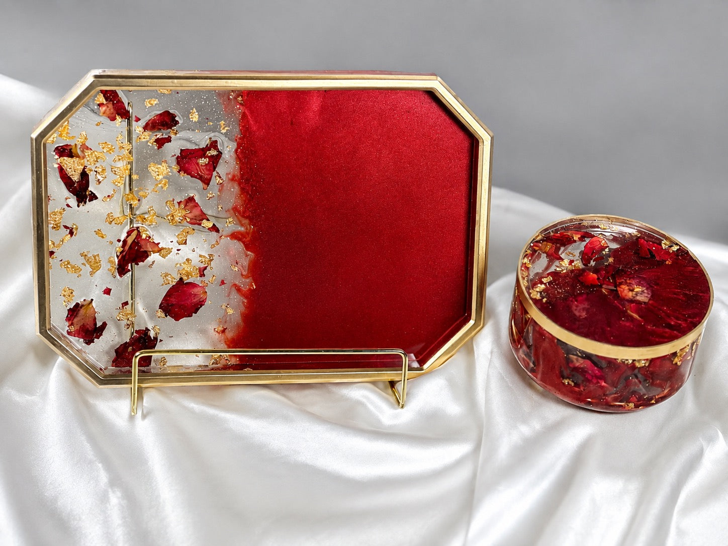 A decorative octagonal tray with a gold-tone frame and a round matching lidded box, both filled with red resin, dried red flower petals, and gold leaf, displayed on white satin fabric against a gray background.