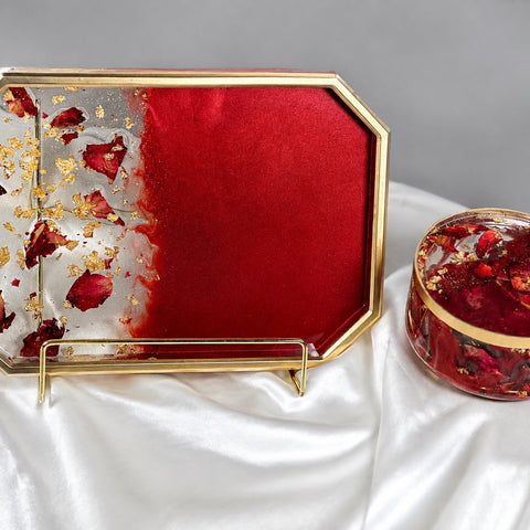 A decorative octagonal tray with a gold-tone frame and a round matching lidded box, both filled with red resin, dried red flower petals, and gold leaf, displayed on white satin fabric against a gray background.