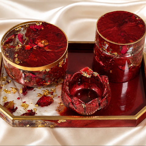 Three red resin decorative containers with gold trim and dried flower inclusions, displayed on a matching red and gold tray over a cream satin fabric background.