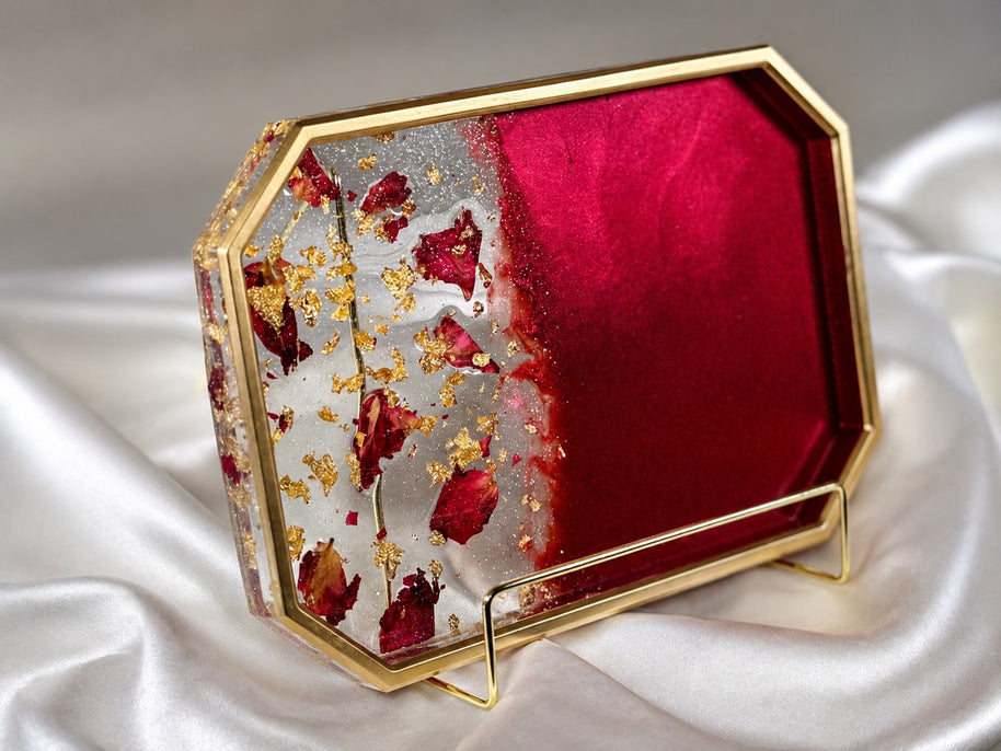 An octagonal decorative tray with a gold metal frame, featuring a split design of deep red velvet on one side and a clear side filled with dried red rose petals and gold leaf flakes on the other. The tray is displayed on a white satin fabric background.