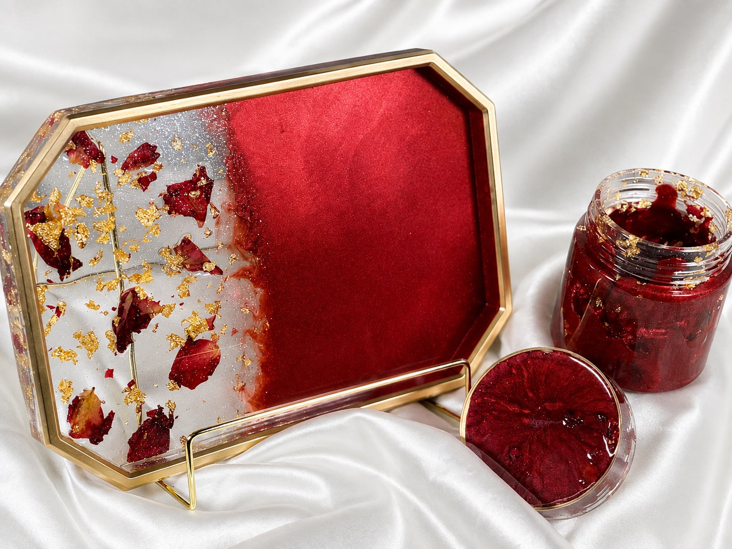 A decorative octagonal tray with a gold frame and a translucent red-and-white design, shown alongside a glass jar and lid containing a red preserve or jam, all arranged on white satin fabric.