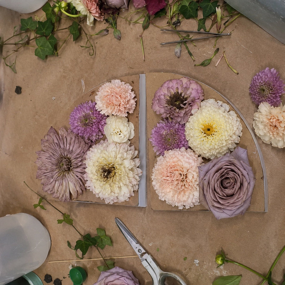 Two clear acrylic flower boards filled with dried and fresh flowers, including mauve roses, purple and cream dahlia-like blooms, arranged on a brown paper work surface with scissors and floral scraps nearby.