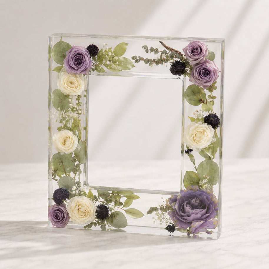 A clear acrylic picture frame decorated with dried purple and cream roses, green leaves, and small black thistle flowers, standing on a light marble surface against a white background.