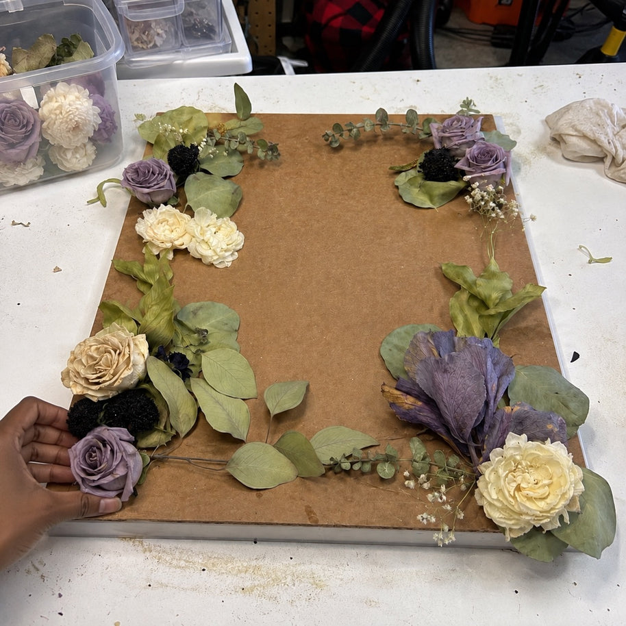 A hand is arranging a border of dried flowers and greenery on a square brown board, set on a white worktable. The floral arrangement includes cream roses, lavender-purple roses, pale yellow flowers, and green eucalyptus leaves.