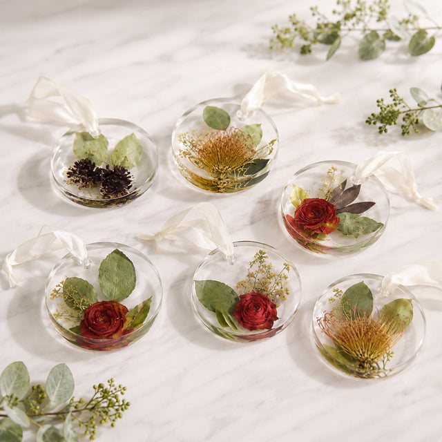 Six round clear glass ornaments with white hanging ribbons, each containing pressed flowers and green leaves, arranged on a light marble surface with small floral sprigs around them.
