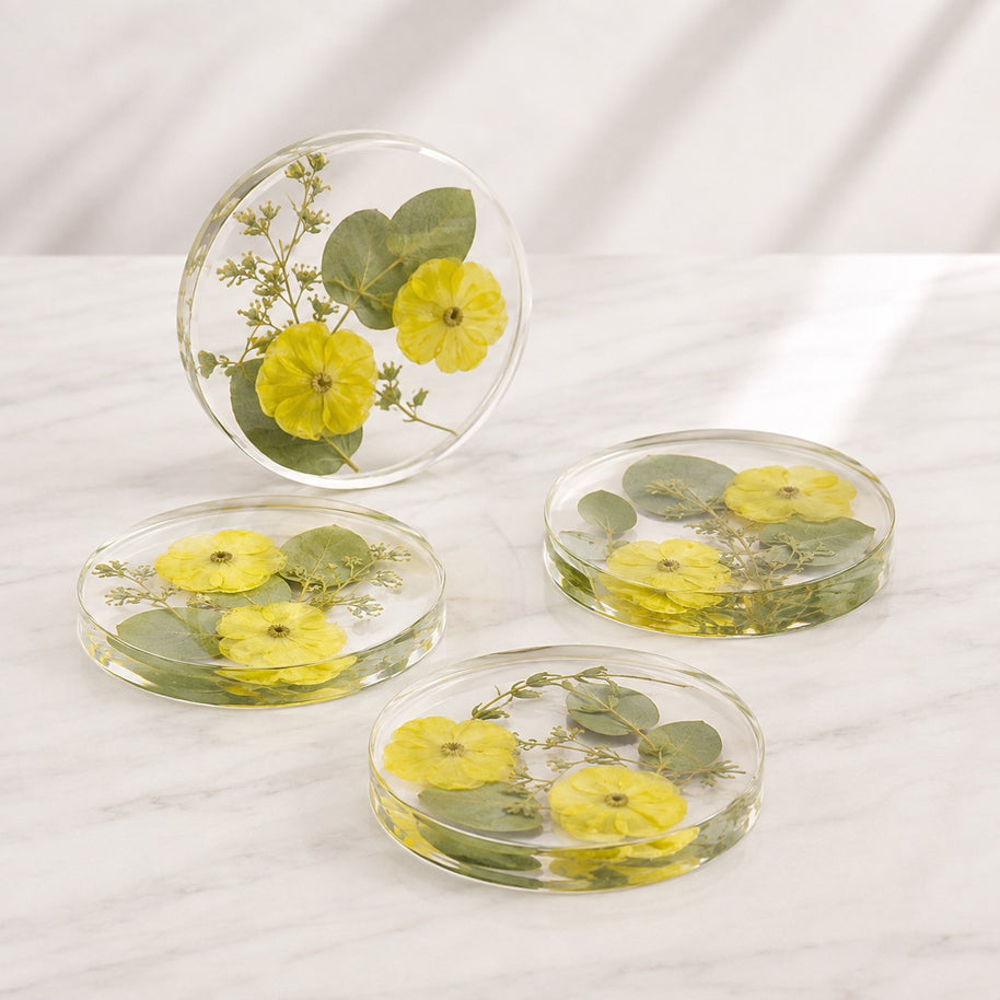 Four round clear resin coasters with pressed yellow flowers and green leaves, arranged on a white marble surface with one coaster standing upright.