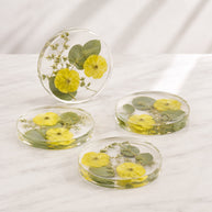 Four round clear resin coasters with pressed yellow flowers and green leaves, arranged on a white marble surface with one coaster standing upright.
